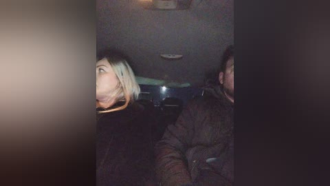 Video of two people in a dimly lit car. A blonde woman with short hair in a black jacket gazes left, while a man in a dark jacket looks forward.