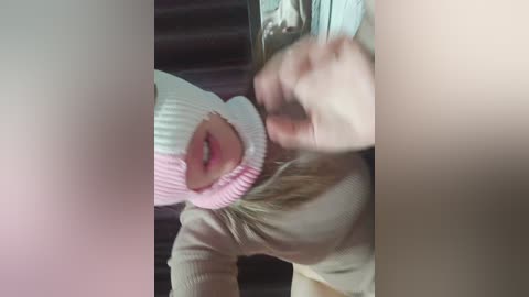 A video of a young child with fair skin, wearing a white knitted hat and beige sweater, smiling playfully, captured from a low angle. The background features a dark, possibly wooden, surface.