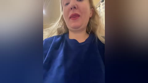 Video of a smiling, fair-skinned woman with blonde hair, wearing a blue medical gown, lying on a hospital bed with blurred background.