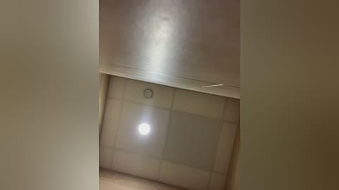 Video of a dimly lit, beige-tiled ceiling with a circular light fixture in the center, creating a soft, diffused glow. The walls are painted a light cream color, and the floor is not visible.