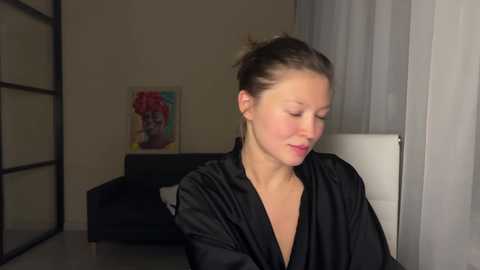 Video of a fair-skinned woman with brown hair tied in a bun, wearing a black satin robe, sitting in a dimly lit room with a black couch, a colorful painting, and sheer curtains.