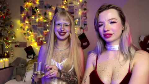 Video of two smiling women in festive, low-light setting; one blonde, one brunette, both wearing revealing outfits, holding wine glasses.