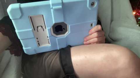 Video of a light-skinned person holding a light blue, iPad-like device with a black camera lens and a visible hinge. The person is wearing a black top and is sitting in a beige car seat.