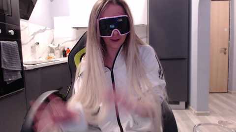 Video of a blonde woman wearing a virtual reality headset, seated in a modern kitchen. She has long hair, a white jacket, and appears to be holding a glass.
