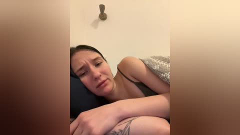 Video of a woman with long dark hair, wearing a black bra, lying on a bed with a gray blanket, looking distressed, with a visible tattoo on her arm.