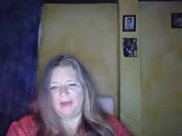 Video of a middle-aged Caucasian woman with long, light brown hair, wearing a red shirt, standing in a dimly lit room with a yellow wall and a framed picture.