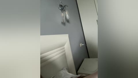Video of a minimalist bedroom with a white tufted headboard, gray walls, a small bedside table, and a wall-mounted sconce with glass shades. The room is dimly lit, creating a serene and understated atmosphere.
