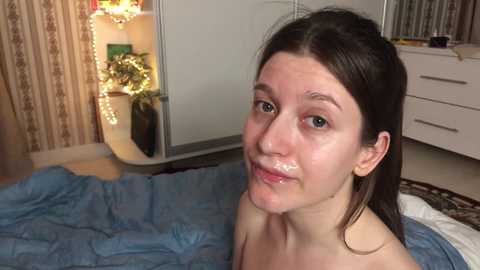 A video of a young woman with long dark hair, lying on a bed with a blue blanket, in a room with a white wardrobe, a shelf with plants, and a dresser.