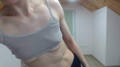 A video of a slender, fair-skinned woman with a flat chest wearing a light gray ribbed crop top, standing in a dimly lit attic with white walls and wooden beams.