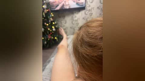 Video of a woman with light brown hair lying on a couch, watching a TV with a Christmas tree and a framed picture in the background.