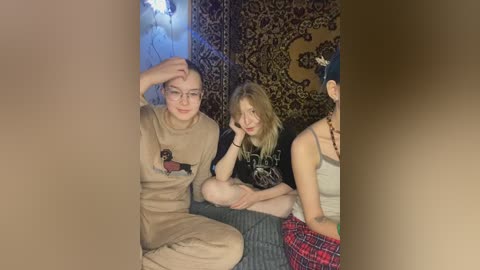 Video of three young people in a dimly lit room with a leopard print tapestry in the background. The young man on the left wears a beige sweatshirt and pants, the young woman in the center has blonde hair and a black graphic tee, and the young woman on the right wears a red plaid skirt.
