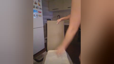 A video of a naked woman standing in a modern kitchen, holding a sheet of paper. The kitchen has white cabinets, a refrigerator adorned with colorful magnets, and a tiled backsplash.