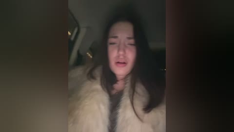 Video of a young woman with long dark hair, wearing a fur coat, crying with a distressed expression. The background is dimly lit, with blurred objects, possibly in a home setting.