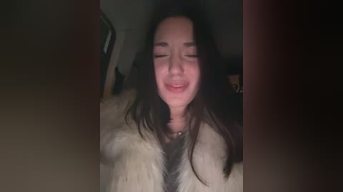 A low-resolution video of a smiling Asian woman with long black hair, wearing a white fur coat, taken in dim lighting.
