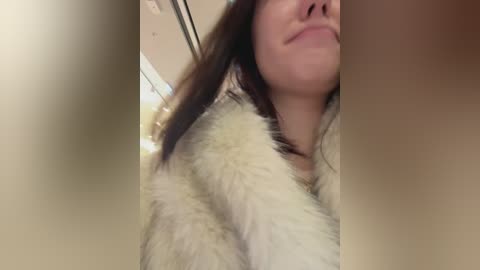Video of a close-up, partially blurred face of a woman with fair skin, brown hair, and a faint smile, wearing a white, fluffy coat. Background features a beige wall.
