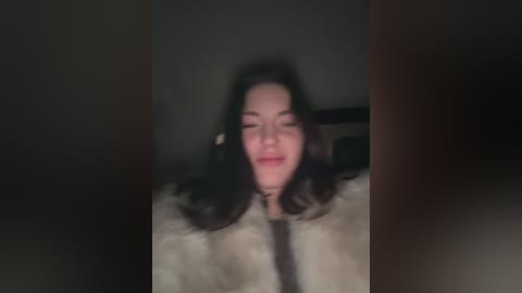 A dimly lit video of a young woman with long black hair, wearing a fur coat, captured in a blurry, night-time indoor setting.