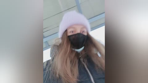 Video of a woman with long brown hair, wearing a light pink beanie, black mask, and dark jacket with a fur collar, standing indoors against a blurred background.