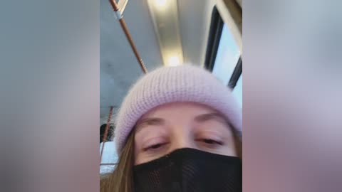 Video of a woman with light skin, wearing a light pink knit beanie, black face mask, and a black jacket, taken from a low angle inside a subway car.
