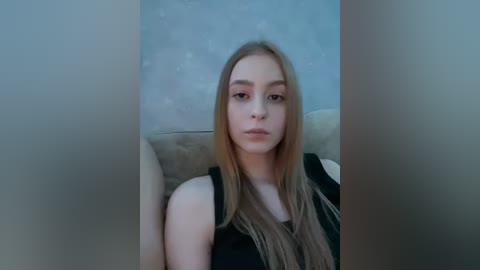 A video of a young Caucasian woman with long, straight blonde hair, wearing a black tank top, sitting on a beige sofa with a muted blue wallpaper background.