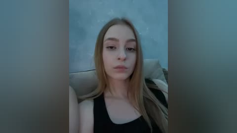 Video of a young Caucasian woman with long, straight blonde hair, wearing a black tank top and beige jacket, sitting on a beige couch in a dimly lit room with a blue wall.