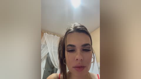 Video of a young woman with olive skin and brown hair, wearing a red top, looking down in a dimly lit room with a white canopy bed in the background.