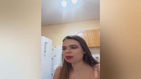 A video of a woman with long brown hair, wearing red lipstick and a pink top, standing in a beige-walled kitchen with wooden cabinets and a white refrigerator.