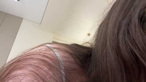 Video of a close-up view of a person's back and shoulder, showing a dark brown, straight-haired woman with a silver hair extension. The background features a beige wall and part of a white ceiling.