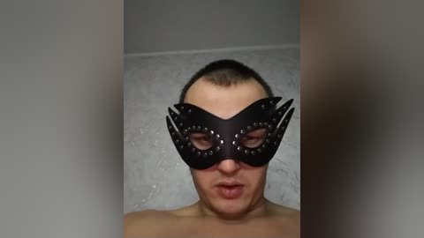 Video of a man with short brown hair, wearing a black masquerade mask with silver studs, standing against a gray textured wall, looking forward.