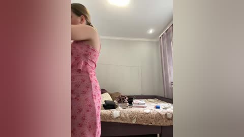Video of a woman in a pink floral dress standing in a dimly lit bedroom, with a bed covered in a patterned quilt and a small dog lying on it.