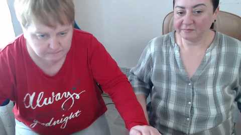 Video of two middle-aged women with fair skin, one in a red shirt, the other in a gray plaid shirt, sitting closely on a bed, holding hands, in a simple, brightly lit room.