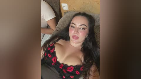 Video of a young woman with long black hair, fair skin, and medium breasts, wearing a revealing black top with red lips. She is reclining on a brown couch, with a man partially visible in the background.