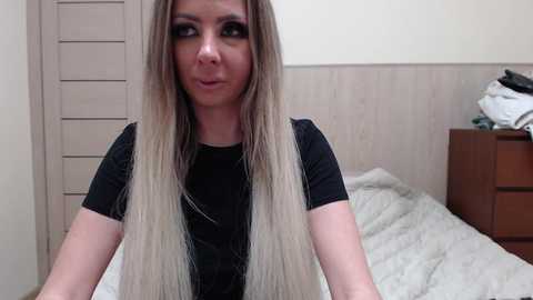 Video of a young woman with long, straight blonde hair, wearing a black top, standing in a bedroom with white bedding, wooden furniture, and a closed shutter window.