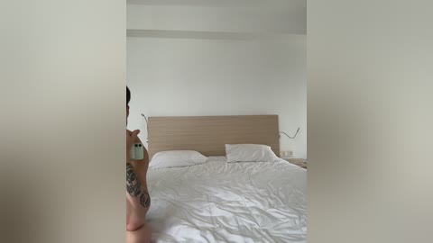 Video of a minimalist bedroom with a light wooden headboard, white bed linens, and a person with tattoos taking a selfie with a phone.