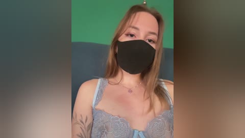 Video of a fair-skinned woman with long, light brown hair, wearing a blue lace bra, black face mask, and a green background.