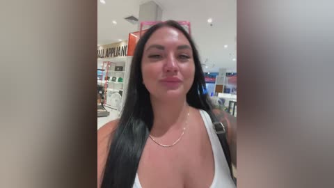 Video of a smiling, fair-skinned woman with long black hair, wearing a white tank top, standing in a brightly lit drugstore with shelves in the background.