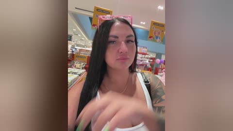 A video of a young woman with long black hair, wearing a white tank top, holding a pink drink can, taken in a brightly lit, modern store with colorful displays and shelves in the background.