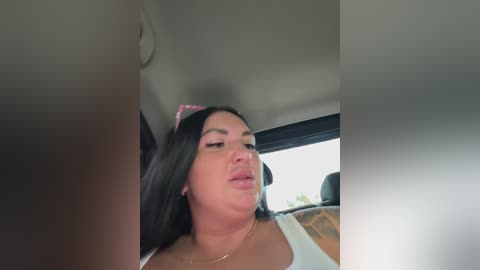 Video of a woman with long black hair and a pink hairband, wearing a white tank top, sitting in a car. She has a soft, slightly pouty expression.