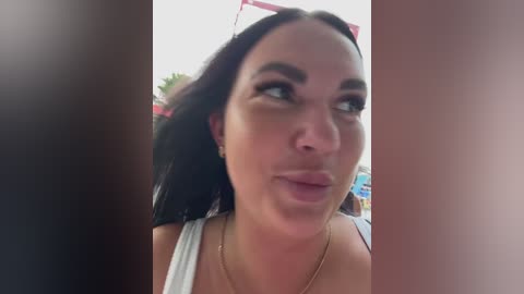 Video of a woman with dark hair, wearing a white tank top, making a pouty face, with blurred background.