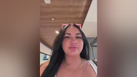 Video of a young woman with long black hair, wearing a white tank top, standing in a modern, wood-paneled room. She has light skin and a neutral expression.