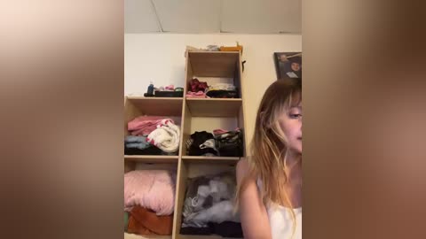 Video of a young woman with long, blonde hair, wearing a white towel, standing in a cluttered bedroom with beige walls and wooden shelves filled with clothes and personal items.