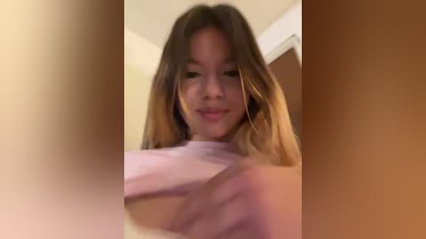 A video of a young woman with light brown hair, wearing a light pink top, smiling while looking at the camera. The background is blurred, featuring beige walls.