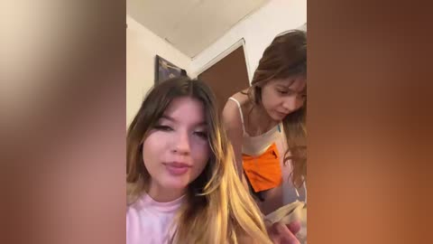Video of two young women in a bedroom. The woman on the left has long, blonde hair, a pink shirt, and a confident expression. The woman on the right, with long brown hair, a orange top, and a worried look, is adjusting her top.