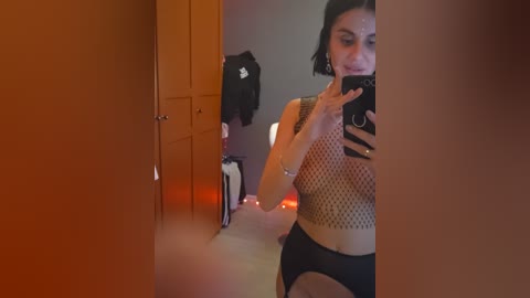 A video of a woman with short black hair taking a mirror selfie in a dimly lit room. She is wearing a black fishnet top and black underwear, with wooden cabinets and a black jacket hanging in the background.