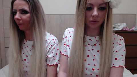 Video of a blonde woman with long hair, wearing a white t-shirt with red heart patterns, in a dimly lit bedroom with wooden furniture and a bed.