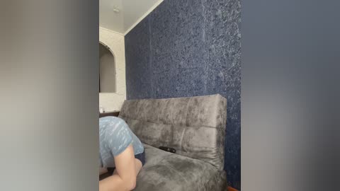 A video of a person in a light blue shirt and dark shorts kneeling on a gray couch against a blue textured wall, with a white arched doorway in the background.