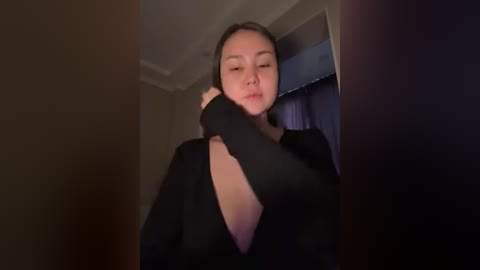 A video of an Asian woman with long black hair, wearing a black long-sleeved top, leaning against a door frame in a dimly lit room.