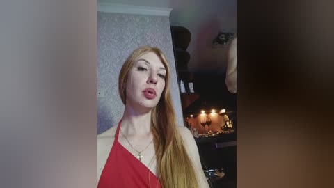 Video of a fair-skinned, red-haired woman with a slim figure, wearing a red halter top, posing indoors. Background features a patterned wallpaper and a lit chandelier.