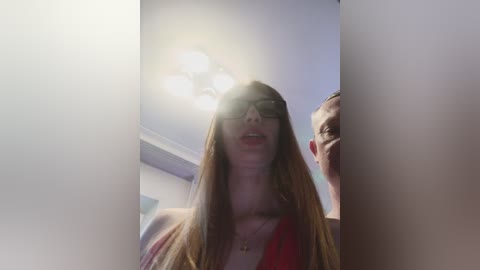 A video of a young Caucasian woman with long, straight brown hair, wearing black sunglasses, a red dress, and a necklace, standing in a brightly lit room with a chandelier and another person partially visible in the background.