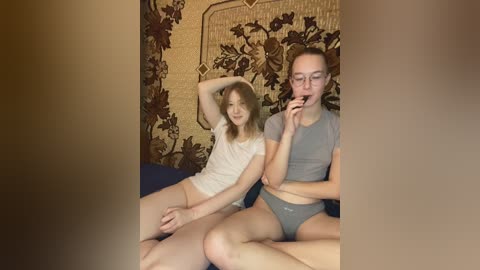 Video of two young women with fair skin and light hair, sitting on a bed, wearing casual gray shirts and underwear. One chews gum, the other rests her arm. Background features floral-patterned wallpaper.