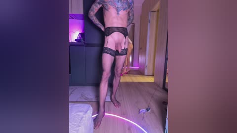 Video of a muscular, tattooed man with pale skin wearing black lace garter belt and stockings, standing in a dimly lit modern bedroom with wooden floors, lit purple neon ring, and visible electrical cords.